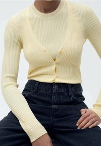 Hellgelber gerippter Cardigan mit tiefem V-Ausschnitt und Knöpfen, darüber ein passendes geripptes Oberteil, kombiniert mit hoch taillierten schwarzen Jeans.