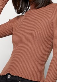 Maglione a collo alto a coste in marrone smorzato, con design slim-fit e orlo scallop. La texture morbida e le maniche lunghe creano una silhouette aderente.