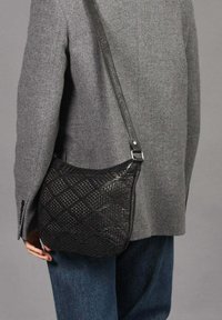 Bolso de hombro de cuero trenzado negro con un patrón diagonal, que cuenta con una correa larga y un borde superior suave. Llevado sobre un abrigo gris.