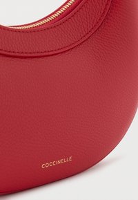 Borsa a mano in pelle rossa con superficie testurizzata e forma arrotondata. Presenta una cerniera dorata e un logo impresso in oro sul fondo.