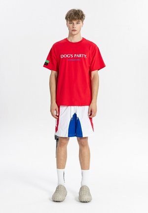 Joven de pie mirando hacia adelante con camiseta roja "DOG'S PARTY", pantalones cortos blancos y azules, calcetines blancos y zapatos beige texturizados sobre un fondo blanco liso.