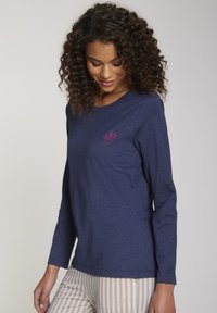 Vrouw met krullend haar, gekleed in een marineblauwe blouse met lange mouwen en een klein roze bloemmotief op de borst, en lichte gestreepte broek.