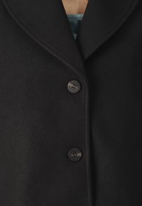 Černý vlněný kabát s rozparkem na klopě, vybavený dvěma kulatými černými knoflíky, měkkou texturou a střihem na tělo. Detailní pohled na horní část.