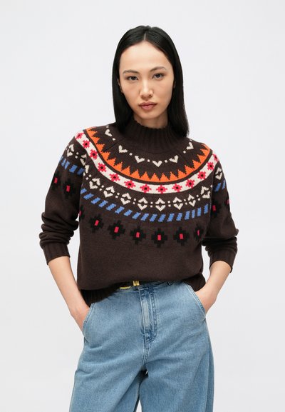 Naine, kes kannab tumepruuni kampsunit erksavärviliste geomeetriliste mustritega ja helesiniseid kõrge vöökohaga teksaseid, seisab käed taskutes.