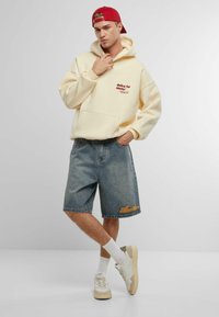 Felpa gialla con testo rosso, pantaloni corti di jeans e sneakers bianche. Dettagli notevoli includono una tasca frontale e un design ricamato sui pantaloni.