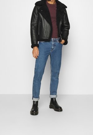 Schwarze Lederjacke mit Lammfellfutter, kastanienrotes Hemd, selbstgefaltete blaue Jeans und schwarze Knöchelstiefel. Insgesamt lässiges Outfit.