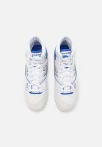 Paire de baskets montantes New Balance 650 blanches et bleues vues de dessus sur un fond clair uni.