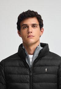 Sort pufferjakke med høj krave og vandret quiltning, med en sølvlogo accenter på venstre bryst og et gråt indre lag.