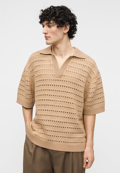 Νέος άνδρας με σγουρά μαλλιά, φορώντας μπεζ πουλόβερ με ανοιχτή πλέξη και κοντά μανίκια, και καφέ παντελόνι, κοιτάζει προς τα αριστερά του.
