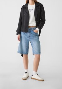 Zwarte trenchcoat, witte sweatshirt met logo, lichtblauwe denimshorts, witte sneakers met zwarte accenten, gedragen met witte sokken.