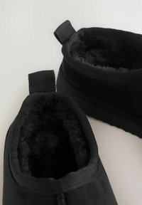 Svarta tofflor i mjukt mocka med ett plyschigt, lurvigt inre. Har dragflikar för enkel påtagning och texturerade gummisulor.