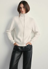 Biała bluzka na zamek błyskawiczny z prążkowaną teksturą i wysokim kołnierzem, zestawiona z czarnymi skórzanymi spodniami. Bluzka ma pionowe detale i długie rękawy.