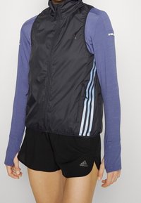 Impermeabile nero senza maniche con collo alto, tasche con cerniera e strisce Adidas blu lungo i lati; abbinato a una maglia a maniche lunghe blu con trama.