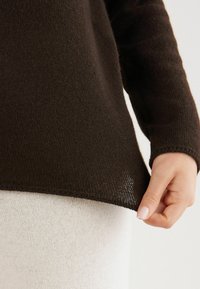 Dunkelbrauner gestrickter Pullover mit weitem Schnitt, strukturierter Stoff und gerippten Bündchen. Hellfarbiger Stoff darunter sichtbar. Hand hält den Saum.