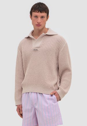 Młody mężczyzna w beżowym, teksturowanym swetrze z kołnierzem i pastelowo różowych szortach w paski, stoi z jedną ręką w kieszeni.