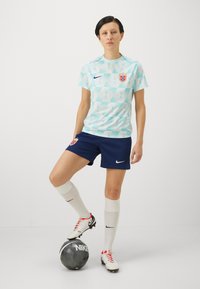 Nike kvinnefotballsett med en lyseblå skjorte med geometrisk mønster, marineblå shorts, hvite sokker og rød-acenterte fotballsko, sammen med en svart ball.