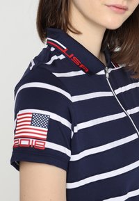 Polo-shirt bleu marine avec des rayures blanches, avec un col zippé, du texte rouge "USA" et une patch drapeau avec "2018" sur la manche.