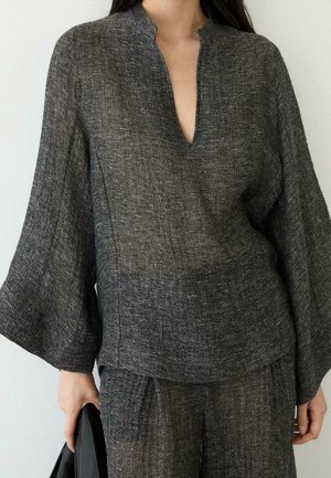 Femme portant un haut ample à manches longues texturé gris foncé avec un décolleté en V profond et un pantalon large assorti, tenant un objet noir dans une main.