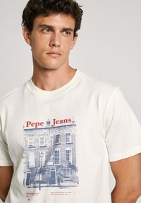 Krémové tričko s modrou a červenou grafikou budovy, obsahující text "Pepe Jeans" a ilustrovaný detail lampy na ulici.