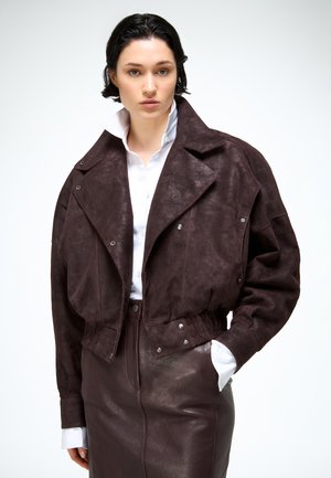 Femme aux cheveux courts et foncés portant une veste surdimensionnée en daim marron foncé et une jupe en cuir assortie, avec une chemise blanche à col en dessous.