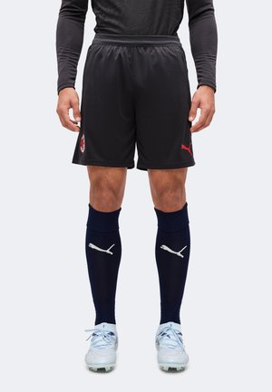 Homme portant un short de football noir avec les logos AC Milan et Puma, des chaussettes Puma bleu marine et des chaussures à crampons blanches, debout sur fond blanc.