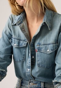 Ανοιχτό μπλε πουκάμισο από τζιν με κουμπιά πέρλες, δύο τσέπες στο στήθος και κλασικό γιακά. Φθαρμένη υφή με διακριτική ξεθώριασμα.