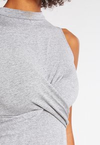 Harmaa hihaton toppi, jossa korkea kaulus ja vyötäröllä kiertävä yksityiskohta. Pehmeä kangas ja sileä pinta.