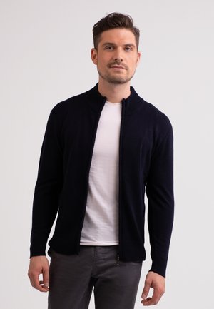 Suéter azul marino con cremallera, cuello de canalé, textura de punto y diseño ajustado; llevado sobre una camiseta blanca y combinado con pantalones grises.