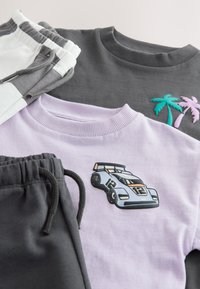 To sæt børnetrøjer og joggingbukser; en grå trøje med palmebladspatches, en lavendel trøje med et racerbilsbadge, kombineret med grå og hvide bukser.