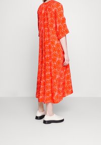 Robe maxi rouge à motifs avec tissu semi-transparent, coupe décontractée, manches trois-quarts et silhouette fluide, associée à des chaussures blanches.