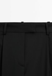 Close-up van de tailleband van zwarte op maat gemaakte broek met riemlussen en verborgen voorkeursluiting.