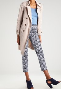 Ljusbeige trench coat med bruna knappar, blå top, mönstrade marinblå byxor och marinblå klackskor. Skräddarsydd, figurnära silhuett.