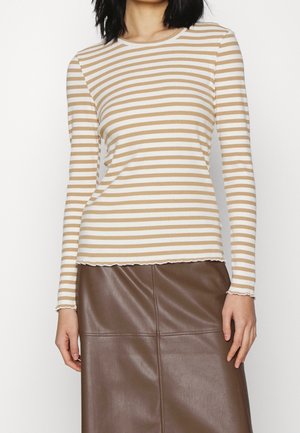 Femme portant un t-shirt à manches longues beige et blanc à rayures horizontales, associé à une jupe en cuir marron.