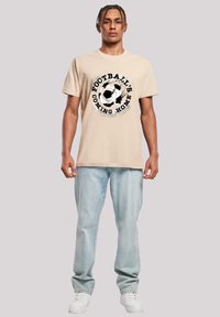 Beige cotton t-shirt featuring a black and white soccer ball graphic with text “FOOTBALL’S COMING HOME,” paired with light blue denim jeans and white sneakers.