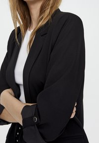 Black blazer with a tailored fit and folded sleeves. Constructed from smooth fabric, featuring a subtle sheen and button accents.