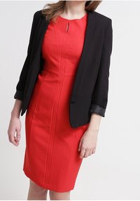 Rode jurk met een aansluitende silhouet, voorzien van verticale naden en een keyhole-detail, gecombineerd met een zwarte blazer met opgerolde mouwen.