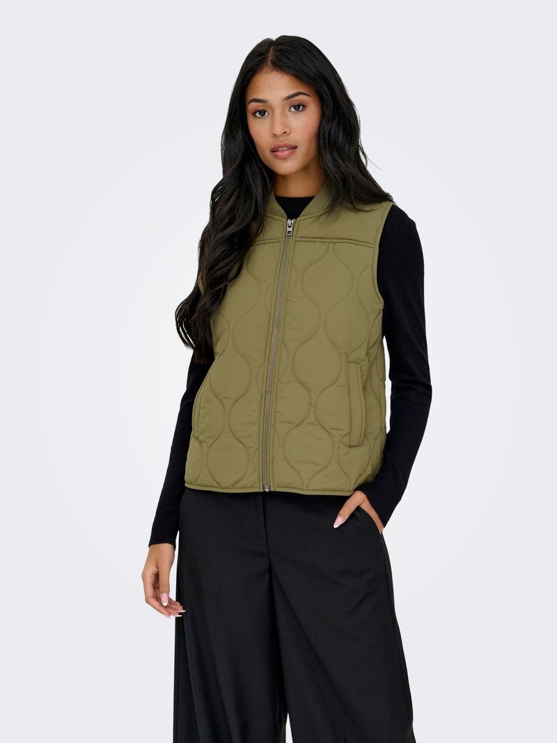 Femme aux longs cheveux foncés portant une veste matelassée kaki zippée par-dessus un haut noir à manches longues et un pantalon noir à jambes larges, debout avec une main dans la poche.