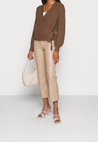 Pull marron côtelé à enfiler avec ceinture nouée à la taille, pantalon en cuir synthétique beige, crop top blanc, sac à main blanc texturé, et sandales à talons blanches à lanières.