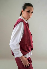 Rode textuurvest over een wit shirt, met een ontspannen pasvorm en zijzakken. Het model draagt een paarse broek en een gouden ring.