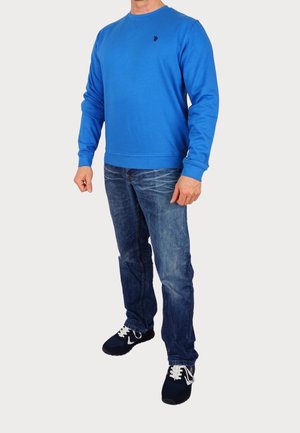 Sudadera azul con cuello redondo y puños de canalé, combinada con jeans azul desgastados y zapatillas negras con cordones blancos. Detalle de logotipo bordado simple.