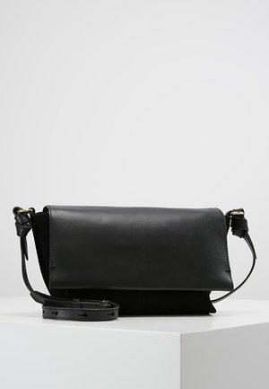 Sort læder firkantet crossbody taske med justerbar rem og guldfarvet spænde, hvilende på hvid overflade mod ensfarvet baggrund.