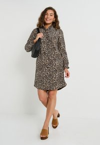 Vestido de estampado de leopardo en tela ligera con cuello, mangas largas y un corte holgado, combinado con mules de tacón marrones y un bolso negro.