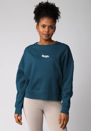 Vrouw met krullend haar die een teal sweatshirt met de tekst "Nope." en beige ribgebreide legging draagt, staande tegen een effen lichte achtergrond.