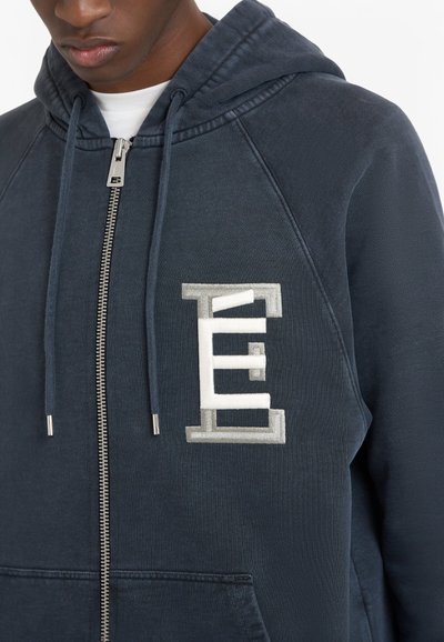Sweatshirt à capuche bleu marine avec fermeture éclair, doté d'un tissu texturé, d'une poche kangourou et d'un accent logo blanc et argent sur la poitrine.