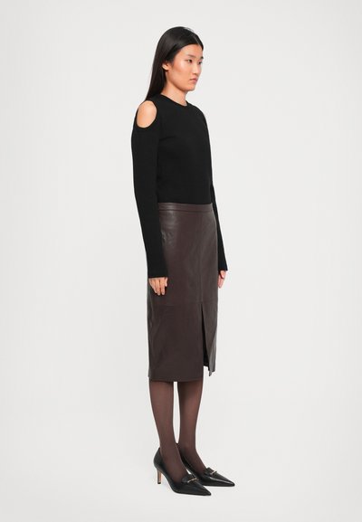 Woman in black cold-shoulder sweater, dark brown leather midi skirt with front slit, sheer dark tights, and black pointed heels with gold accents.