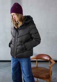 Sort pufferjakke med hætte, med diagonale stikninger og trykknapper. Bæres sammen med blå jeans og en rippelavet lilla hue. Brun stol i baggrunden.