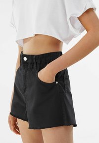 Shorts de mezclilla negra con cintura alta, dobladillo deshilachado, bolsillos frontales y detalle en botón plateado. Combinados con una camiseta blanca corta.
