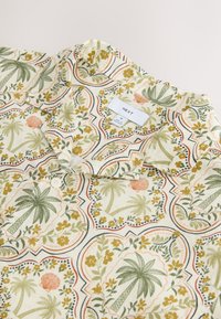 Chemise beige à col avec motifs de palmiers verts et fleurs jaunes, avec une étiquette blanche de la marque NEXT et un bouton blanc visible.