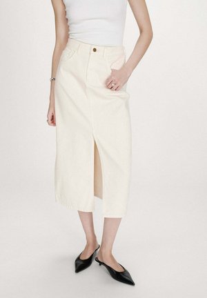 Femme portant un haut blanc sans manches, une jupe midi couleur crème avec une fente à l'avant, et des escarpins noirs à bout pointu avec bride arrière, debout sur un fond blanc.