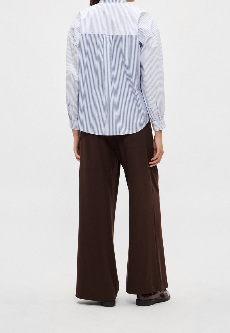 Personne portant une chemise à manches longues rayée bleue et blanche et un pantalon marron ample, debout de dos.